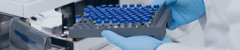 Gloved hands holding a tray of sample vials being loaded into a laboratory analytical instrument, used for impurities testing in pharmaceutical analysis.