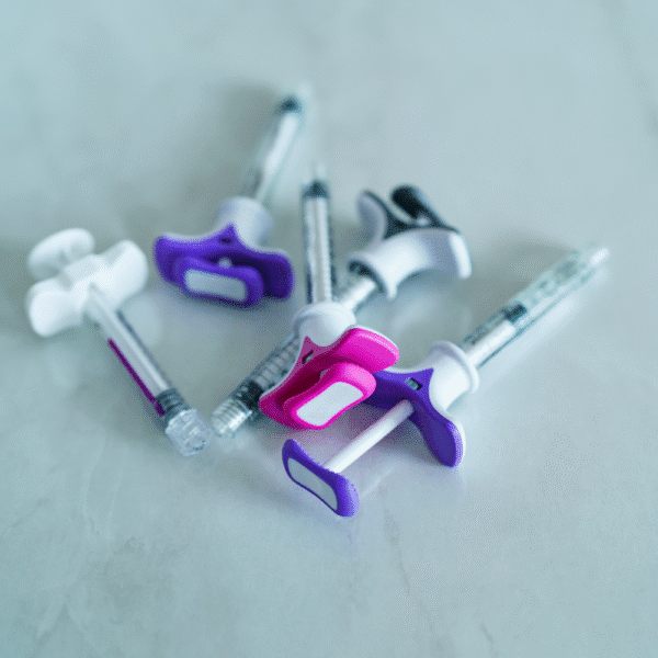 Close-up studio image of multiple colourful pre-filled syringes with safety plunger grips on a clean clinical surface.