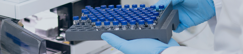Gloved hands holding a tray of sample vials being loaded into a laboratory analytical instrument, used for impurities testing in pharmaceutical analysis.