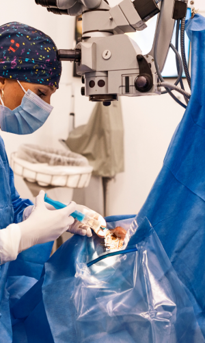 Close-up photograph of an ophthalmic surgeon performing eye surgery under a surgical microscope in a clinical operating room.