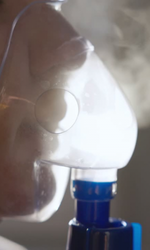 Person using a nebulizer mask connected to a respiratory therapy device, delivering inhaled medication as visible mist.